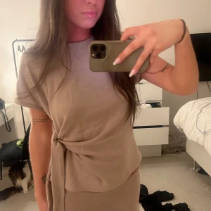 Beige stickad topp med knyt - Säljer en beige topp som tillhör ett set med kort ärm och snygg knytdetalj i midjan. Den har en enkel och stilren design med rak passform och mjukt material. Aldrig använd. Storlek M men passar mig som alltid har S 