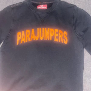 Svart sweatshirt från Parajumpers - Svart sweatshirt från Parajumpers med orange logga framtill och patch på ena axeln. Tröjan har rund halsringning och långa ärmar. Perfekt för dig som gillar streetwear och vill ha något bekvämt och snyggt.