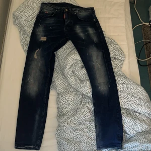 Mörkblå slitna jeans från Dsquared2 - Säljer ett par mörkblå jeans från Dsquared2 med slitningar och färgstänk för en cool, använd look. Klassisk femficksmodell med raka ben och tydlig logga bak i midjan. Perfekta för dig som gillar streetstyle.