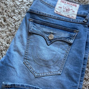 True Religion Ricky Relaxed Straight jeans - Säljer ett par blå True Religion Ricky Relaxed Straight jeans med klassiska kontrastsömmar och stora bakfickor med lock och knapp. Jeansen har en rak passform och tydliga detaljer på fickorna. Perfekta för dig som gillar streetwear och y2k. Skulle säga den passar S och M eftersom midjan är gjort för män och är stor. 