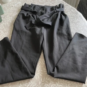 Svarta kostymbyxor med bälte - Snygga svarta kostymbyxor med tillhörande bälte i midjan. Byxorna har normal passform och raka ben, perfekta för en stilren look. Stängs med knapp och dragkedja framtill.