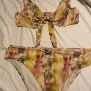Färgglad bikini med volang och knyt - Säljer en bikini med volangdetaljer och knyt framtill. Färgen är en mix av rosa, gul, grön och beige i ett abstrakt mönster. Överdelen har breda axelband och underdelen är klassisk. Perfekt för stranden! Bikini i tie-dye mönster med en knut i mitten av överdelen.  Underdel 50 överdel 95 D