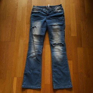Blå bootcut jeans från Rainbow - Säljer ett par blå bootcut jeans från Rainbow med slitningar och hål på båda benen. Jeansen har låg midja.