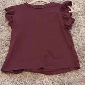 Vinröd topp med volangärmar - Säljer en vinröd topp från Gina Tricot med snygga volangdetaljer på ärmarna. Storlek 146/152. Den är knappt använd, endast 1 gång.