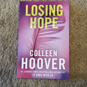 Losing Hope (oanvänd) - Säljer boken 'Losing Hope' av Colleen Hoover. Omslaget är lila med en fjäder och tydlig gul och vit text. Pocketbok på engelska, utgiven av Simon & Schuster. Perfekt för dig som gillar samtida romaner och gripande berättelser.