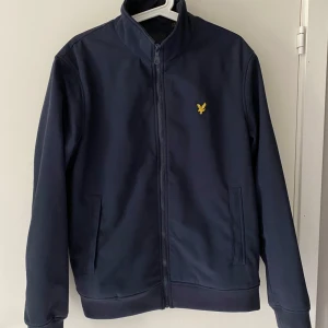 Mörkblå jacka från Lyle & Scott - Säljer en stilren mörkblå jacka från Lyle & Scott med gul logga på bröstet. Jackan har dragkedja framtill, ståkrage och två fickor. Perfekt för dig som vill ha en enkel och snygg look.