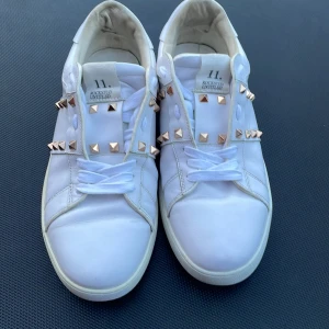 Vita Valentino Rockstud sneakers - Säljer ett par vita Valentino Rockstud sneakers med ikoniska nitar i guld längs sidorna och bak på sulan. Skorna är i skinn och har en klassisk rund tå och platt sula. Perfekta för dig som vill ha en lyxig och edgy stil. Är osäker på storleken men dem passar mig som har storlek 42/43 så har du det så passar dem. Hör av dig vid frågor