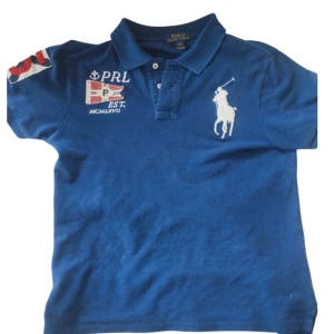 Blå pikétröja från Polo Ralph Lauren - Säljer en blå pikétröja från Polo Ralph Lauren med klassisk krage och knappar. Tröjan har broderad logga på bröstet och detaljer med flagga och text på ena sidan. Perfekt för en sportig och stilren look. Barn L = XS