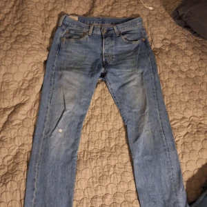 Blå Levis 501 jeans - Klassiska blå Levis 501 jeans med raka ben och femficksmodell. Byxorna har knappgylf och en liten sliten detalj på vänster ben samt bakficka. Perfekta för en avslappnad stil.