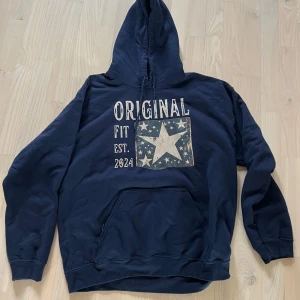 Mörkblå hoodie  - Säljer en mörkblå hoodie med stor vit stjärna och texten 'ORIGINAL FIT EST. 202A' på bröstet. Tröjan har huva och magficka, samt långa ärmar. Perfekt för en avslappnad stil. Storlek L.