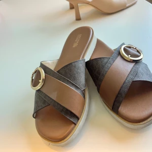 Bruna sandaler med gulddetalj från Michael Kors - Snygga sandaler med breda korslagda remmar i brunt och mörkgrått. Använda 2-3 gånger, storlek 37. 