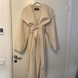 Beige kappa med bälte från Bianca x Nelly - Säljer en stilren beige kappa från Bianca x Nelly i storlek 34. Passar även storlek 36. Kappan har lång ärm, bred krage och knyts med bälte i midjan. Perfekt för vår och höst när du vill ha en klassisk look. 