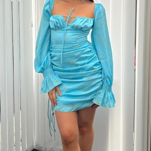 Blå kortklänning med blommigt mönster och volangärmar - Supersöt ljusblå kortklänning med små gula blommor, långa volangärmar och knytning framtill. Klänningen har en figurnära passform och rynkade detaljer längs sidorna. Perfekt för sommarens alla tillfällen! Från ADOORE