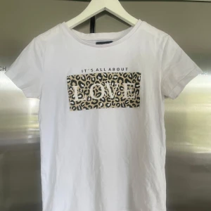 Vit t-shirt med leopardmönster och text - Vit t-shirt med rund halsringning och korta ärmar. Framtill finns ett tryck med leopardmönster och texten 'IT'S ALL ABOUT LOVE'. Perfekt för en trendig och avslappnad stil. Nästan oandvänd, priset går att diskutera💕