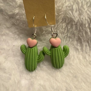 Kaktusörhängen med hjärtan - Säljer ett par unika örhängen formade som gröna kaktusar med söta rosa hjärtan på toppen. Perfekta för dig som vill sticka ut med roliga accessoarer! Hängande modell med krok. Krokarna är i sterling silver 