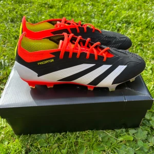Adidas Predator fotbollsskor - Säljer ett par Adidas Predator FG Elit fotbollsskor i svart med vita och röda detaljer samt gula inslag vid hälen. Skorna har snörning och mönstrad ovansida för bättre bollkontroll. Perfekta för spel på gräsplan. Endast provade 1ggr. Säljes pga för liten storlek. Nypris i butik 2800kr