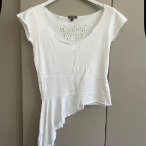 Vit asymmetrisk topp med spetsdetalj från Sisters - Säljer en vit topp från Sisters med asymmetrisk nederkant och spetsdetalj i form av en fjäril fram och bak vid halsringningen. Toppen har korta ärmar och är tillverkad i ett mjukt material. Perfekt för dig som vill ha något unikt och stilrent.