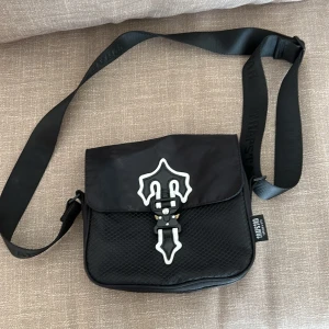 Svart axelväska från trapstar - Cool svart axelväska med justerbart band och unik vit detalj på locket. Väskan har ett spänne framtill och är tillverkad i syntetmaterial med en lätt strukturerad yta. Perfekt för att bära det viktigaste i vardagen.