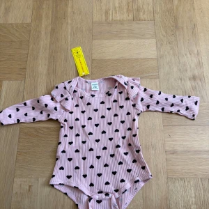 Rosa ribbad body med svarta hjärtan  - Supersöt rosa ribbad body med svarta hjärtan och volangdetaljer vid axlarna. Långärmad modell med knappar i grenen för enkel påklädning. Perfekt för små trendsetters! 3-6 månader oanvänd. Ingår även ett matchande hårband 