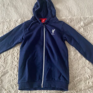 Blå hoodie med dragkedja från Liverpool FC - Mörkblå hoodie med dragkedja från Liverpool FC. Hoodien har huva, långa ärmar och klubbens logga tryckt på bröstet. Perfekt för unga fotbollsfans som vill visa sitt stöd för laget.