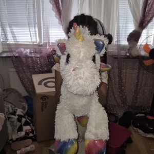 Mjukis Enhörning med regnbågsdetaljer - Säljer en stor, fluffig enhörning i vitt plyschmaterial med regnbågsfärgade hovar, vingar, svans och horn. Den har stora svarta ögon och rosa detaljer på näsan. Perfekt som gosedjur eller dekoration i rummet.