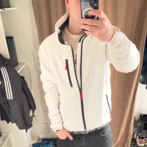 Vit vindjacka från Helly Hansen - Säljer en vit vindjacka från Helly Hansen med svarta och röda detaljer. Jackan har dragkedja framtill, hög krage och flera fickor med dragkedja. HH-logga på bröstet och ärmen. Perfekt för blåsiga dagar.