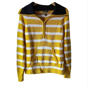 Gul och vit randig tröja med dragkedja från Ralph Lauren - Säljer en gul och vit randig tröja från Ralph Lauren med halv dragkedja och svart krage. Tröjan har långa ärmar, känguruficka och är tillverkad i 100% bomull. Perfekt för dig som gillar färg och klassisk stil!