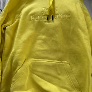 Gul hoodie från Peak Performance - Säljer en gul hoodie från Peak Performance med broderad logga på bröstet. Tröjan har huva med dragsko och en stor magficka framtill. Perfekt för dig som gillar färg och vill sticka ut lite extra.