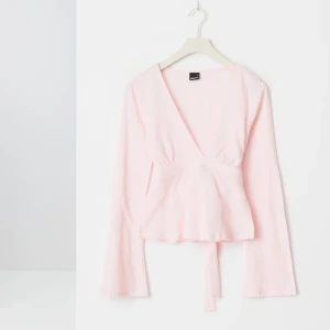 Ljusrosa omlottopp med knytband - Säljer en ljusrosa omlottopp med v-ringning och långa ärmar. Toppen har knytband i midjan som knyts baktill och ger en feminin siluett. Perfekt att styla med jeans eller kjol för en trendig look.