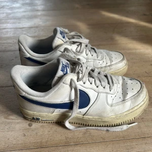 Nike Air Force 1 med blå detaljer - Klassiska Nike Air Force 1 sneakers i vitt med blå swoosh och detaljer. Skorna har snörning, rund tå och platt sula. Perfekta för dig som gillar en tidlös och sportig stil. Ovandelen är i skinn och de har tydlig Nike-logga på baksidan.