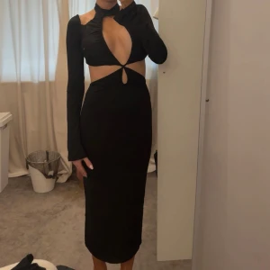 Svart långklänning med cut-out och lång ärm - Säljer en svart långklänning med lång ärm och trendiga cut-out-detaljer framtill. Klänningen har en tight passform, hög hals med choker-effekt och en snygg öppning vid magen. Perfekt för dig som vill sticka ut.