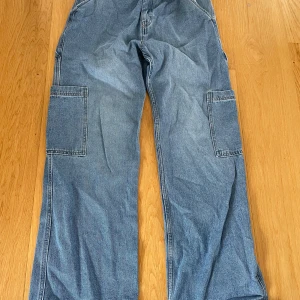 Blå jeans från H&M - Säljer ett par blå jeans från H&M med raka ben och stora fickor på sidorna. Byxorna är tillverkade i jeansmaterial och har en avslappnad passform. Perfekta för en cool och avslappnad stil.