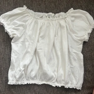Vit topp  - Söt vit offshoulder topp från KappAhl med korta puffärmar och resår vid halsringning och nederkant. Toppen har en lätt rynkad struktur och volangdetaljer längs kanterna. Perfekt för en somrig look!
