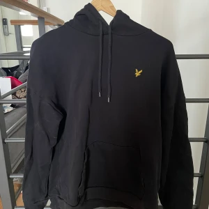 Svart hoodie från Lyle & Scott - Säljer en svart hoodie från Lyle & Scott med klassisk gul logga på bröstet. Tröjan har huva med dragsko och en stor magficka framtill. Tillverkad i 100% ekologisk bomull, perfekt för en avslappnad stil. M passar