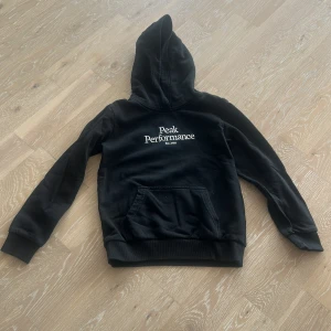 Svart hoodie från Peak Performance - Säljer en svart hoodie från Peak Performance med vit logga på bröstet. Tröjan har huva och en stor magficka framtill. Perfekt för en avslappnad stil och enkel att matcha med det mesta.