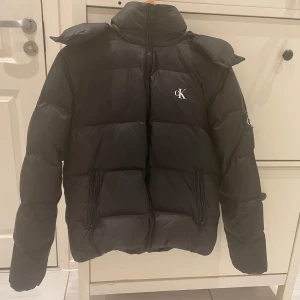 Svart pufferjacka från Calvin Klein Jeans - Säljer en svart pufferjacka från Calvin Klein Jeans med huva och dragkedja. Jackan har två fickor med dragkedja och diskret CK-logga på bröstet. Perfekt för kyliga dagar.