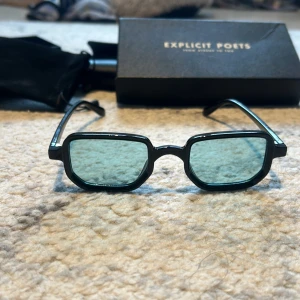  solglasögon från Explicit Poets - Solglasögon med svart, kantig båge och gröntonade glas från Explicit Poets. Glasögonen har en modern, fyrkantig form och levereras med originalfodral.