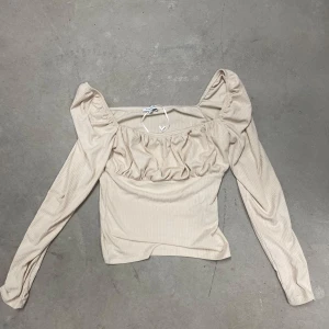 Beige ribbad topp med puffärmar från Bubbleroom - Säljer en beige ribbad topp från Bubbleroom med långa puffärmar och rynkade detaljer framtill. Toppen har en fyrkantig urringning och är tillverkad i ett mjukt, stretchigt material. Perfekt för dig som gillar trendiga detaljer!
