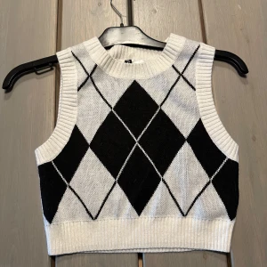 Vit och svart stickad väst med rutor från hm - Säljer en stickad väst från hm i vitt med svarta rutor i argylemönster. Modellen är croppad med bred ribbad kant vid ärmhål, hals och nederkant. Perfekt att styla över en skjorta eller t-shirt.