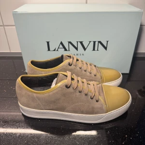 Lanvin sneakers i mocka och skinn - Säljer ett par snygga Lanvin sneakers med beige mocka och olivgröna detaljer i skinn på tån och runt snörningen. Skorna har rund tå, platt vit sula och klassisk snörning. Perfekta för dig som gillar stilrena och exklusiva sneakers. Storlek 7 passar 41/42 ‼️köp nu och få ett par gråa Dondup jeans på vår sida‼️