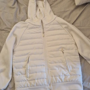 Vit quiltad hoodie med dragkedja - Säljer en vit hoodie med quiltade partier och dragkedja framtill. Jackan har huva, två fickor med dragkedja och långa ärmar. Perfekt för kyliga dagar när du vill ha något stilrent och bekvämt.