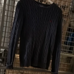 Mörkblå kabelstickad tröja från Ralph Lauren - Säljer en klassisk mörkblå kabelstickad tröja från Ralph Lauren. Tröjan har rund halsringning, långa ärmar och diskret broderad logga på bröstet. Perfekt för en stilren och tidlös look. Lite sliten i kragen