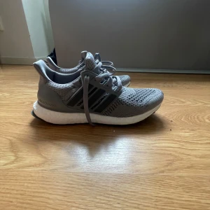 Grå Adidas Ultraboost 20 sneakers - Säljer ett par grå Adidas Ultraboost 20 sneakers med stickad ovandel och vita Boost-sulor. Skorna har klassiska tre ränder på sidan och snörning. Perfekta för både träning och vardag. Märke och modell syns tydligt på taggen.