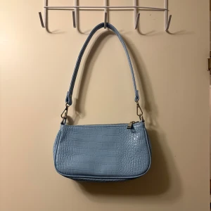 Ljusblå axelväska från Gina Tricot - Säljer en ljusblå axelväska från Gina Tricot med croco-mönstrad yta och dragkedja upptill. Väskan har en enkel, stilren design och ett avtagbart axelband. Perfekt för att lyfta din outfit med en pop av färg.