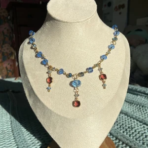 Halsband med blå och röda pärlor - Unikt halsband med blå facetterade pärlor och tre hängande detaljer, varav två röda och en blå. Kedjan är guldfärgad och pärlorna är jämnt fördelade längs hela halsbandet. Perfekt för att ge en färgklick till din outfit.