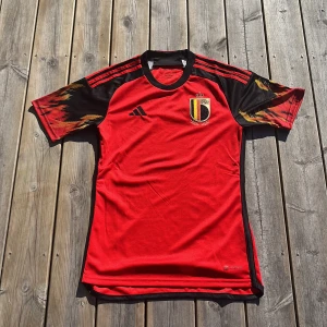 Belgien fotbollströja från Adidas - Säljer en Belgien landslagströja från Adidas i rött med svarta och gula detaljer. Tröjan har korta ärmar med mönstrade partier och broderad landslagssköld på bröstet. Klassisk sportdesign med tre ränder på axlarna. Säsongen 22/23.