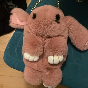 Rosa kaninformad axelväska med kedja - Supergullig axelväska i form av en kanin, gjord i mjuk rosa fuskpäls med vita tassar och svarta ögon. Väskan har en guldfärgad kedja och öppnas med dragkedja upptill. Utrymmet i väskan är inte jättestort men bra om man ska vara ute ett litet tag och  inte behöver så mycket med sig. 