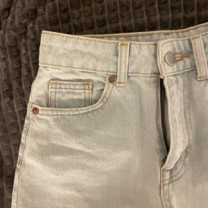 Ljusblå jeansshorts med fransig kant - Säljer ett par ljusblå jeansshorts med hög midja och rå, fransig kant nedtill. Klassisk femficksmodell med knapp och dragkedja framtill. Perfekta för varma dagar och enkel att matcha med olika toppar.