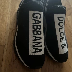 Svarta sneakers från Dolce & Gabbana - Säljer ett par svarta sneakers från Dolce & Gabbana med tydlig logga i vitt på ovansidan. Skorna har en sportig design med elastisk ovandel och kraftig sula med mönster undertill. Perfekta för dig som gillar streetwear och vill sticka ut.