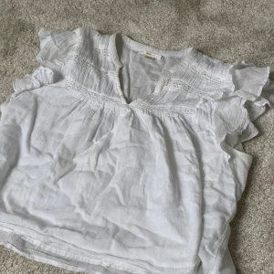 Vit blus med volangdetaljer - Säljer en söt vit blus med volangdetaljer på axlarna och v-ringning. Blusen har fina spetsinslag upptill och är tillverkad i ett lätt och luftigt tyg, perfekt för varmare dagar. I storlek 158/164💗